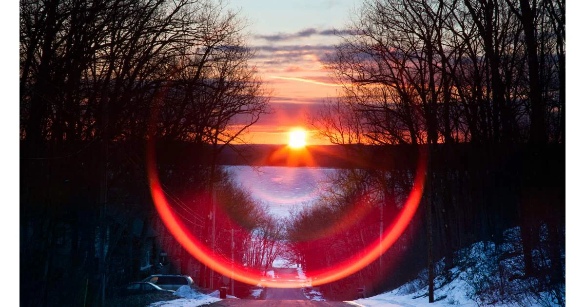 Sunset over a snow-covered road framed by trees, with a prominent circular lens flare in the foreground.
